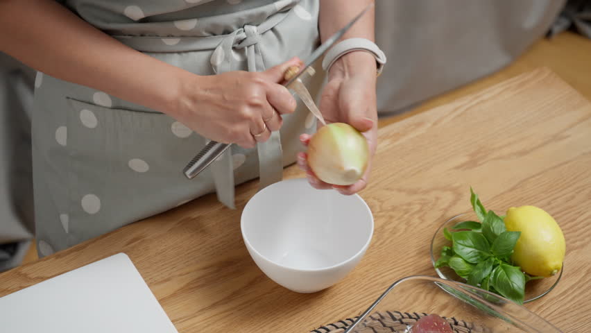 A woman in the kitchen peels the skin from a raw yellow onion with a knife. Preparing fresh vegetable ingredients for a homemade meal. Slicing food for a healthy salad or a vegan dish.