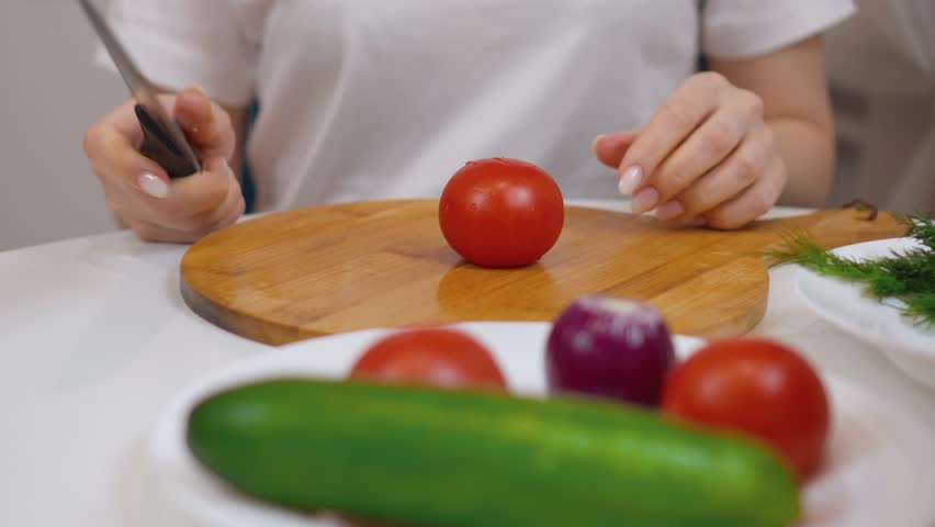 Female chef slices fresh, ripe tomatoes on a wooden cutting board, preparing a healthy and colorful salad with cucumbers, red onions, and dill