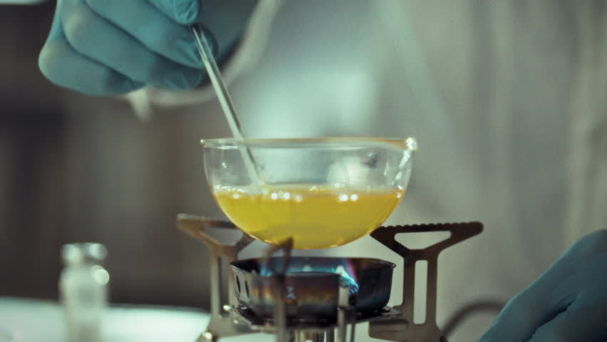 Mixing chemicals in a laboratory experiment using a Bunsen burner for heating. A scientist in gloves stirs a yellow liquid in a glass container on a Bunsen burner - Powered by Shutterstock - Get 15% off with code: PIKWIZARD15