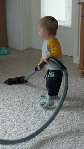 Cute little toddler is standing on a fluffy, soft rug and playfully pushing a vacuum cleaner around, imitating household chores with a sense of curiosity, joy, and fun during playtime