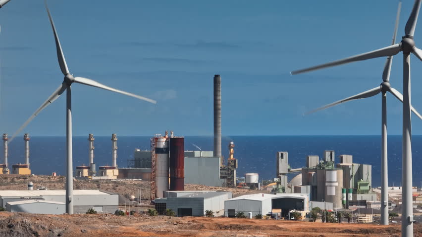Tenerife, Canary Islands: Spinning wind turbines generating clean energy alongside a factory with smoke-emitting chimneys, highlighting Tenerife's energy transition. Drone flight footage