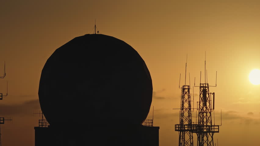 Large spherical radome and communication antenna towers silhouetted against a vibrant orange sunset sky, emphasizing the significance of telecommunications infrastructure. Aerial drone panorama