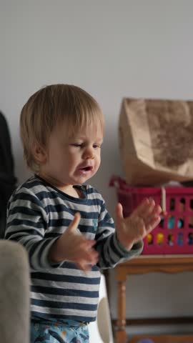 A cute little baby stands thoughtfully in a cozy home environment, clapping his hands with great interest and curiosity. This moment adds warmth and playfulness to his joyful exploration
