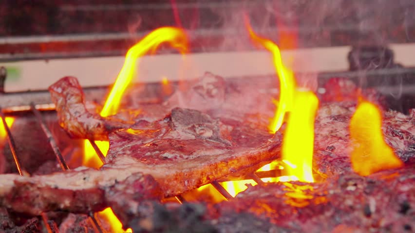 A juicy lamb steak sizzles on a hot barbecue grate. Open flames lick the meat, creating a perfect char and releasing aromatic smoke. The ultimate summer BBQ experience