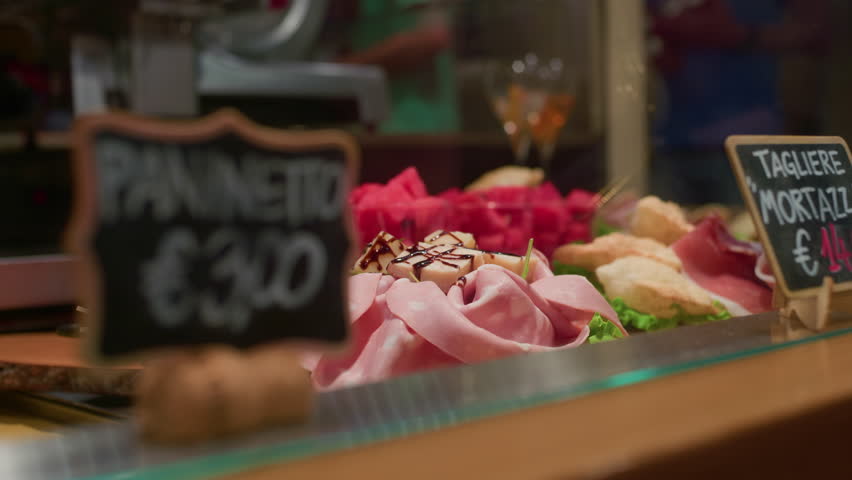 Handheld left-to-right shot of a food stall tray displaying Italian delicacies such as cured meats, cheeses, focaccia, and traditional snacks.