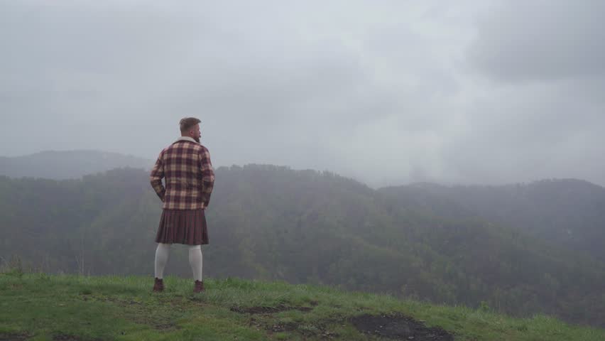 An adult redhead bearded man in Scottish national dress plaid jacket and kilt stands on a hill and looks into the distance. Masculinity, pride and human dignity concept.