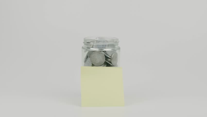 Hand repeatedly inserting coin into full jar with yellow note on white background, symbolizing savings, investment, finance, and money management