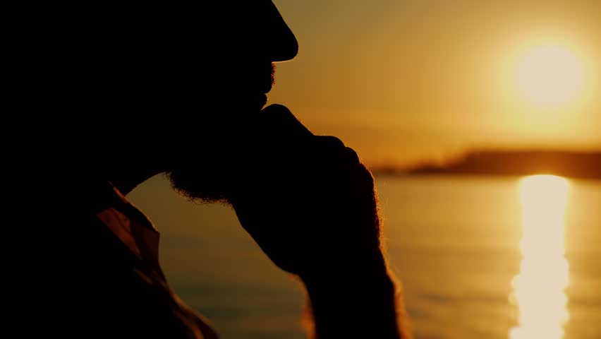 Close-up of the silhouette of a brutal bearded man's face on the ocean shore at sunset. Portrait of a thoughtful guy against the backdrop of the sea. The man brings his hands to his face.