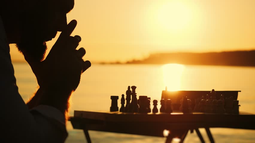 Silhouette of brutal man sitting on seashore at table and playing chess alone at sunset. Lonely man playing chess on the beach.