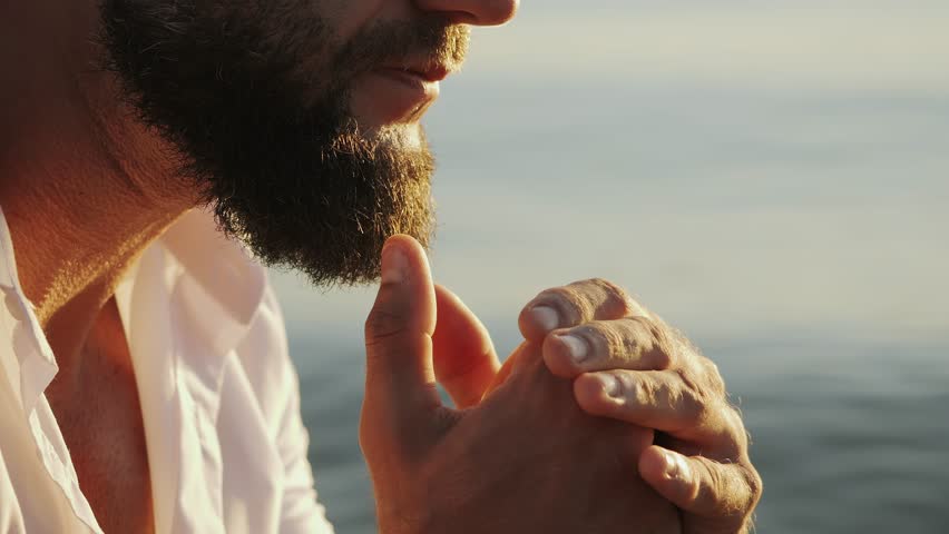 Close-up of a brutal bearded man's face on the ocean shore. Portrait of a thoughtful guy against the backdrop of the sea. The man raises his hands to his face and looks thoughtfully into the distance.