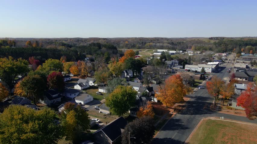 Aerial view of a small Midwestern town in Minnesota during fall. Colorful trees line quiet streets, with residential homes and distant forested hills under clear blue sky.