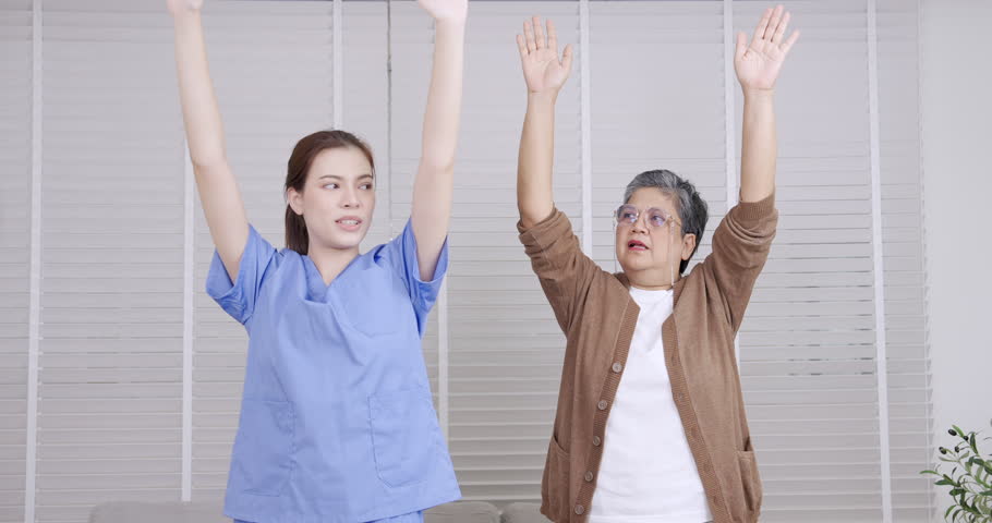 Caucasian young female physiotherapist and Asian senior female patient performing synchronized arm raise exercise in slowmotion session focused on shoulder mobility and balance improvement