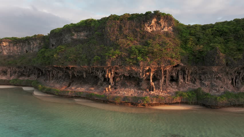 Drone close-up of limestone cliffs in Lekini Bay, Ouvéa, New Caledonia, bathed in golden sunset tones over clear waters.