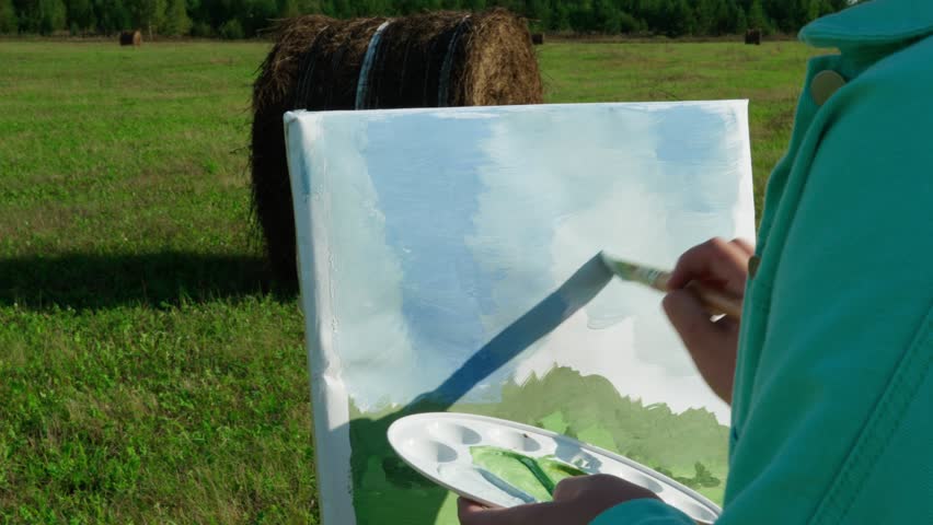 A girl artist paints a landscape in an open field with bales of mown hay. The creative process, the art of inspiration.	