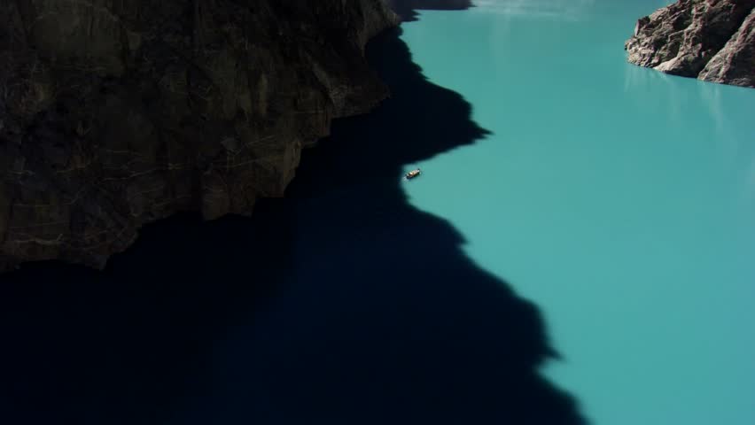 A close-up view of Attabad Lake in Pakistan featuring a small boat on its calm, turquoise waters. The vibrant lake contrasts with steep, rugged cliffs that cast a dark shadow across the surface.