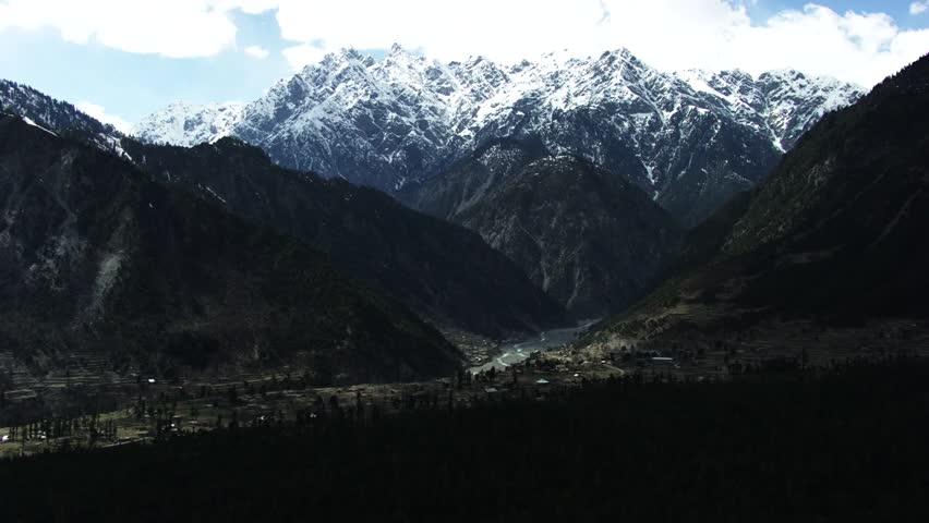 A stunning aerial view of a mountainous valley in Pakistan, featuring snow-capped peaks, dense evergreen forests, and a winding river cutting through terraced fields and a small village.