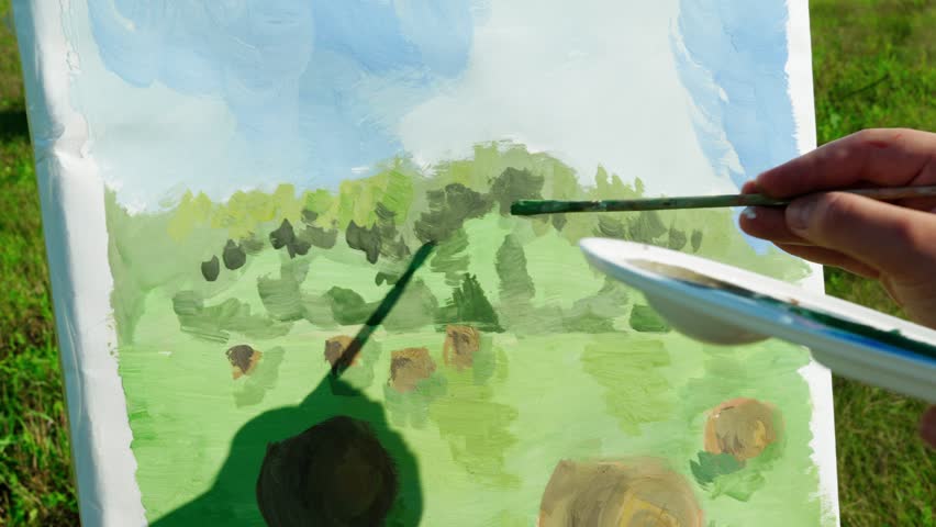 A girl artist paints a landscape in an open field with bales of hay. Close-up of a canvas with a drawing. Creative process, artistic concept.