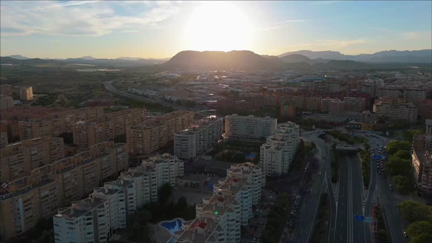 Drone footage pulling back above Alicante city skyline during sunset, showing high-rise buildings and the sun setting behind the mountains in golden light