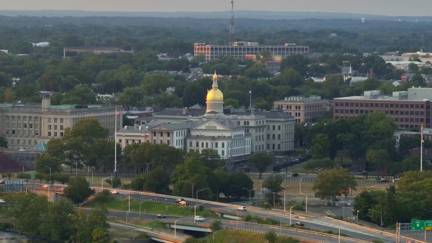 Aerial view of New Jersey State House in Trenton at sunset time in New Jersey. Wide shot. Waving American flag on flagpole. Cars on road along Delaware River.