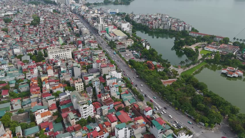 Push-in aerial view of a busy road in Hanoi, Vietnam. A high volume of traffic, including cars and motorbikes, with a mix of buildings and trees.