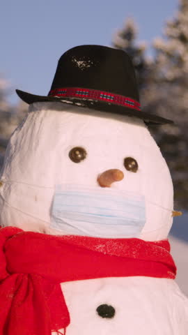 CLOSE UP, DOF: Funny looking snowman wearing a black hat wears a surgical mask to protect him from the coronavirus. Snowman standing in wintry countryside obeys covid19 protocols by wearing a facemask