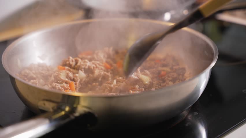 Ground beef is fried with onions and carrot pieces in a pan, while steam rises. A wooden spoon stirs the food during cooking.