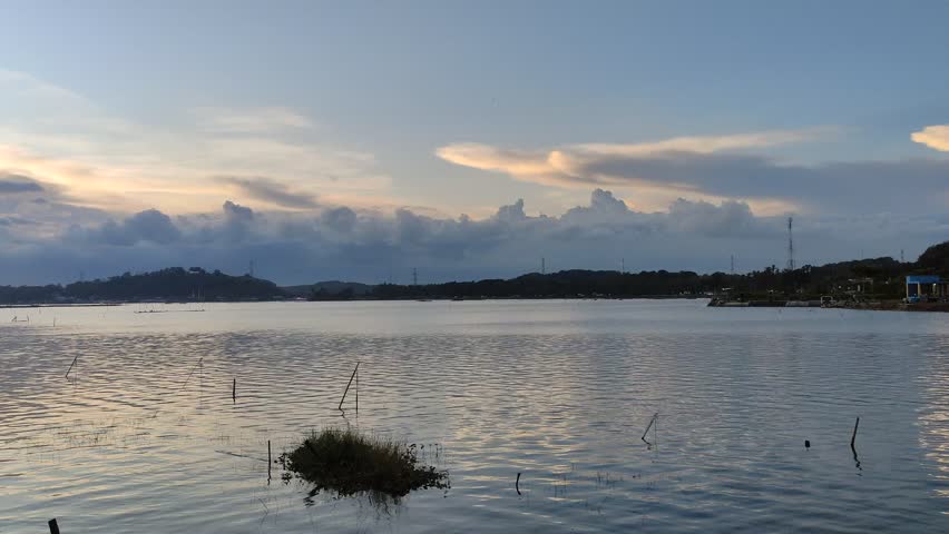 A peaceful view of the lake at dusk, with soft light reflecting on the water. The horizon displays the silhouette of hills against a cloudy sky.