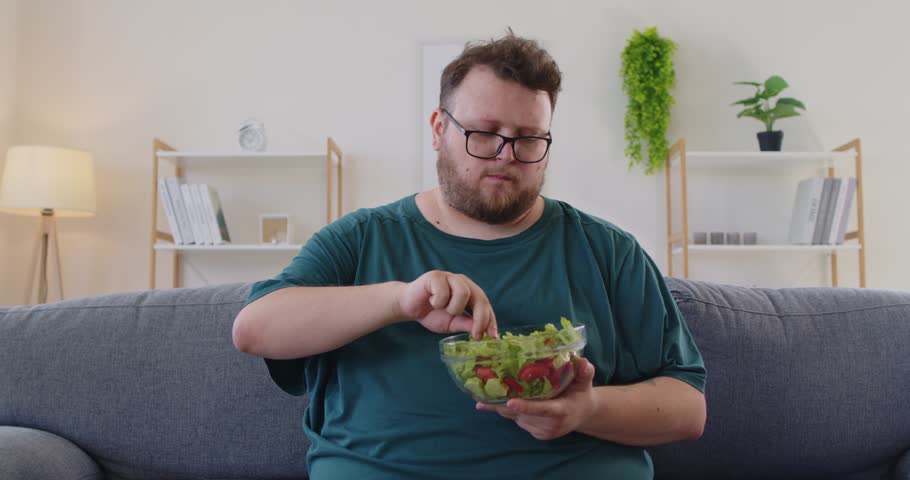 At home, fat overweight man in glasses eats a salad with a fork from a bowl by the window, suggesting a healthy lunch, nutrition and diet in a bright kitchen. Simple healthy meal. Clean life.