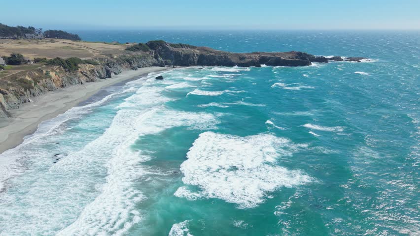 High-altitude drone footage showing California coastline with jetty, large waves, rocky shore, and cliffs during a slight backward movement over the ocean.