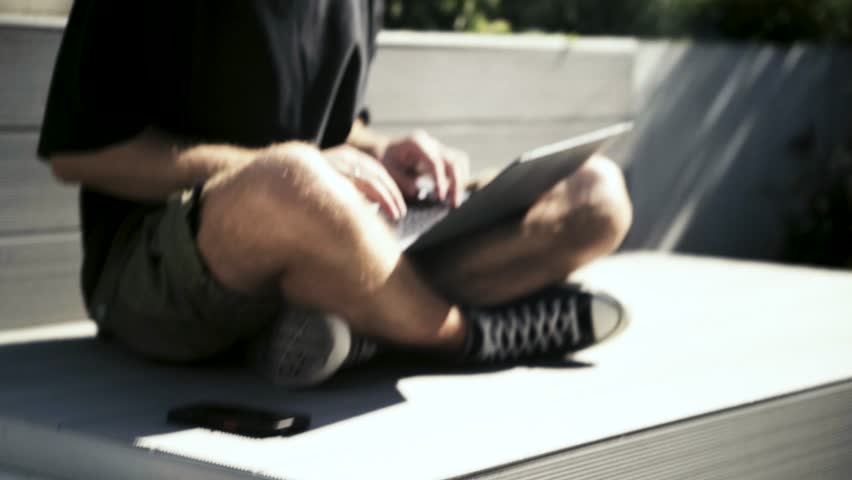 Focused blond freelancer man in black t-shirt typing on laptop keyboard at remote workplace oudoor surrounded by greenery. Portrait of man professional working at laptop remotely on a hot summer day.