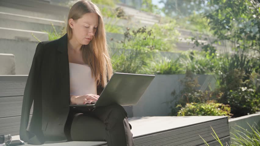 Focused business woman in black suit typing on laptop keyboard at remote workplace oudoors on public steps. Portrait of female professional working at laptop remotely on a hot summer day.
