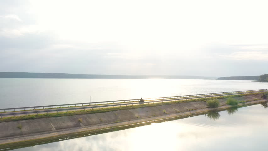 Aerial view to biker riding on motorbike at dam route with sunshine at background. Motorcyclist racing his motorcycle through road near lake. Man drive bike during trip. Concept of adventure. Top view