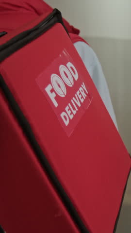 Vertical shot of unrecognizable African American courier going in hallway while carrying big red food delivery bag