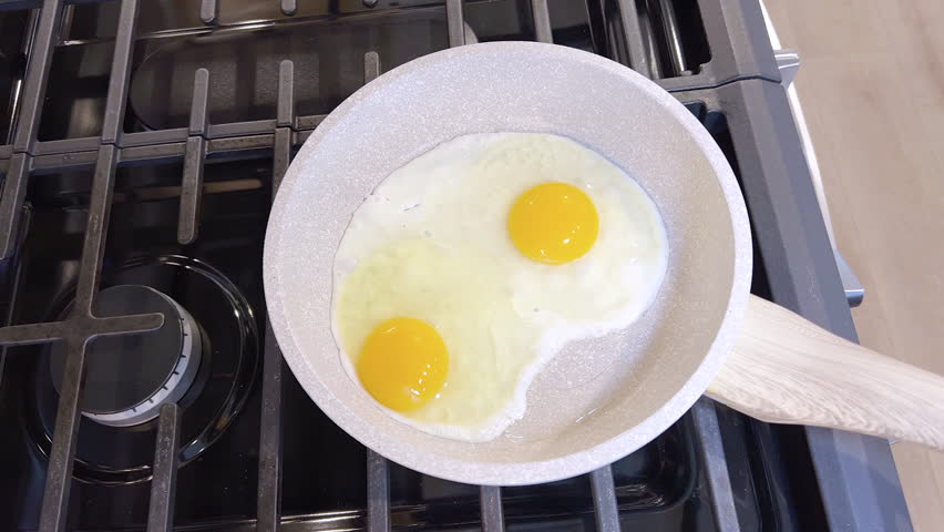 Two perfectly fried sunny side up eggs sit in a non-stick frying pan, with a red silicone spatula ready to serve them, all set on a stove top.