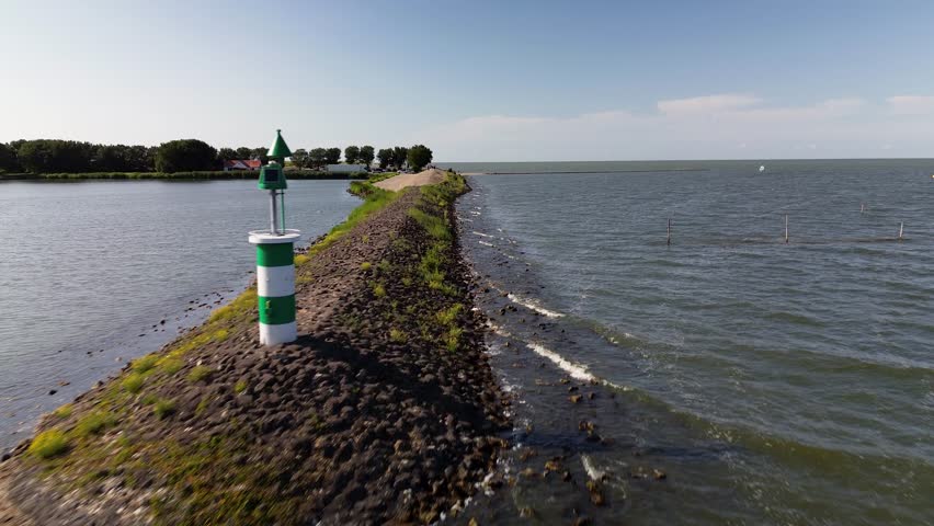 Scenic aerial view of a dam stretching between water bodies, with waves on one side and calm water on the other, surrounded by greenery and a few houses.