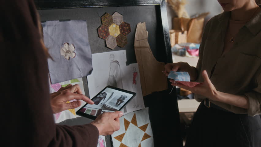 Medium close-up of two female fashion designers choosing fabrics for new garments in workshops, one holding digital tablet with sketches while colleague is suggesting cloth swatches