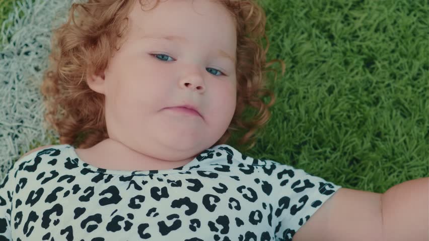 A young girl with curly hair is lying on green grass playfully sticking out her tongue She wears a leopard print shirt and enjoys a carefree moment under the sun