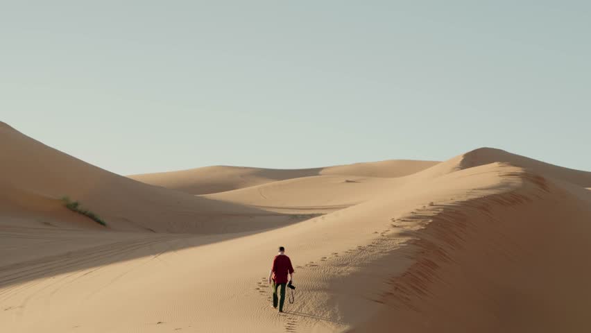 A man walks through the Sahara Desert in Morocco. He is on an adventure and exploring the vast, empty landscape. The sun is setting, creating a beautiful, warm glow. freedom, solitude, wanderlust.