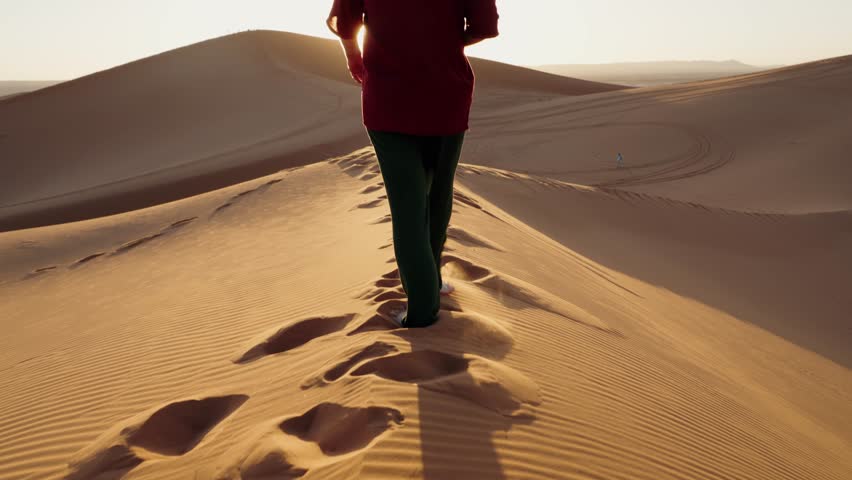 A person walks barefoot across a sand dune in the Moroccan desert at sunset. They leave footprints in the sand as they walk towards the setting sun. In the background, more dunes stretch out.