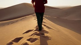 A person walks barefoot across a sand dune in the Moroccan desert at sunset. They leave footprints in the sand as they walk towards the setting sun. In the background, more dunes stretch out. - Powered by Shutterstock - Get 15% off with code: PIKWIZARD15