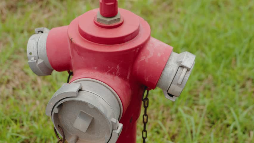 Red fire hydrant standing in green grass field. Close-up shot of the hydrant shows the top part, including the three discharge caps, and the chain attached to the stem.