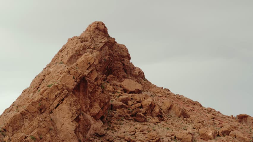 Majestic rock formations in the Moroccan desert landscape. Rugged peaks and canyons create a dramatic scene under a cloudy sky. Orange and brown hues dominate the terrain,