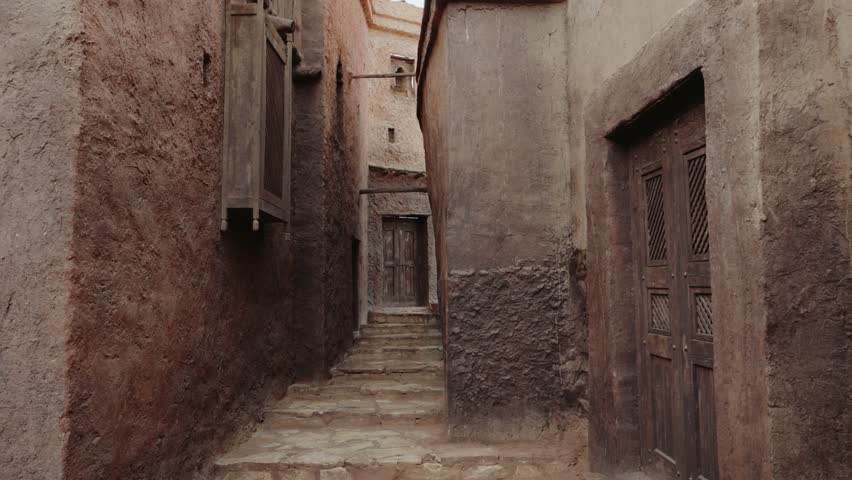 Narrow alley in a Moroccan village with stone steps and wooden doors and window shutters. The walls are made of mud bricks. The scene evokes a sense of history and tradition.