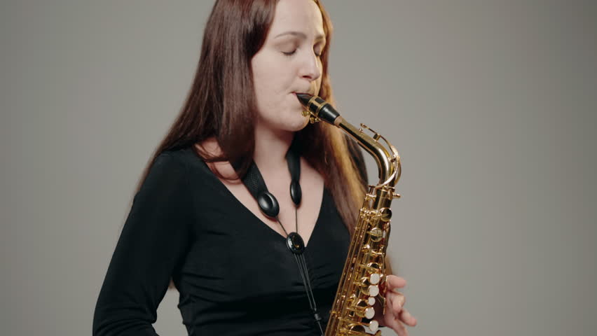 A young woman in black dress plays the saxophone on the white background in studio. A female saxophonist wearing a black evening dress performing a solo on stage.