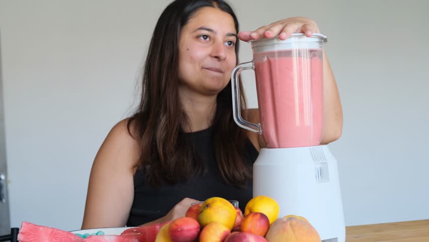 Cute young Middle Eastern woman prepares a watermelon smoothie using a blender. Woman makes a fruit cocktail by mixing watermelon and other fruits. Healthy eating and lifestyle.