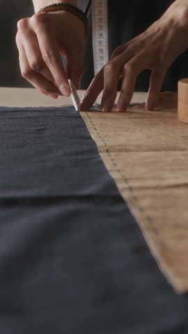Vertical close-up of hands of anonymous woman drawing line with chalk on black cloth around paper template, while preparing to cut fabric for sewing dress in atelier or at home