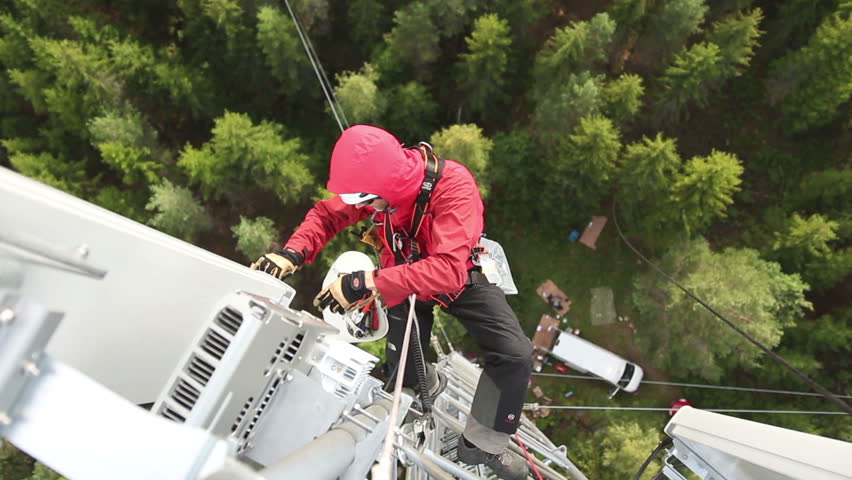 A high-angle view of a telecommunications technician working at height on a telecom tower, performing maintenance on 5G antenna.	