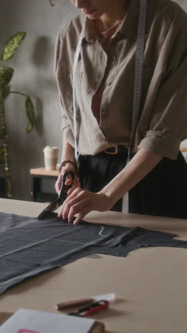 Medium vertical of unrecognizable young seamstress with measure tape carefully cutting black fabric with scissors in atelier, while working on bespoke couture dress design