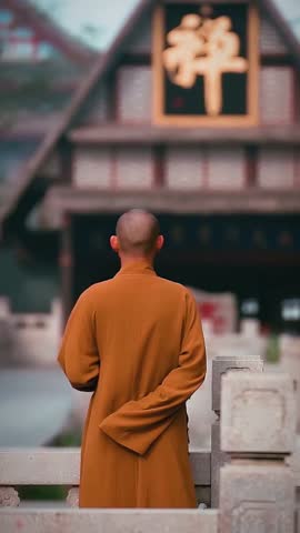 A rear view of a bald monk in traditional robes standing peacefully in front of a temple building with Chinese calligraphy.