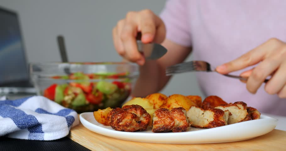 Close up of a woman using a knife and fork, cutting and picking up a piece of meatball from a white plate on a wooden placemat, with a bowl of salad in the background